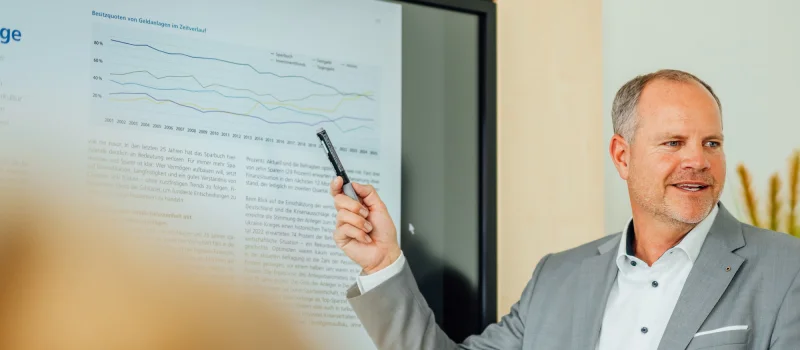 Ein Mann in Geschäftskleidung zeigt mit einem Stift auf eine Präsentation mit Diagramm und Fachtext. Das Bild steht für fachliche Beratung und analytische Entscheidungsunterstützung beim Genossenschaftsverband Weser-Ems.