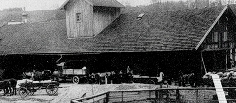Historische Schwarz-Weiß-Aufnahme eines landwirtschaftlichen Gebäudes mit Pferdefuhrwerken und Personen im Außenbereich. Das Bild begleitet den Meilenstein zur Gründung des Vorgängerverbandes des heutigen Genossenschaftsverbands Weser-Ems im Jahr 1890.