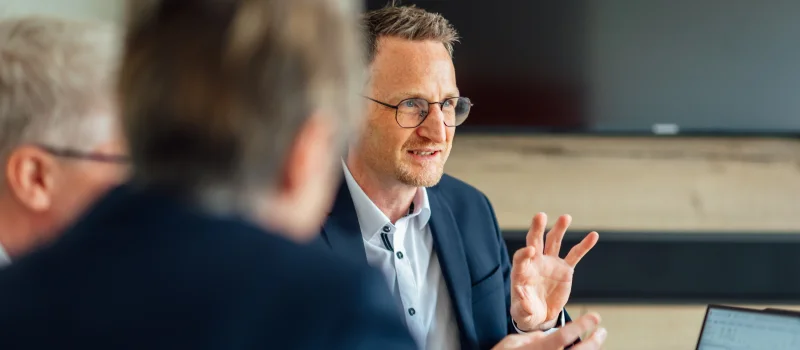 Mehrere Personen sitzen in einem Besprechungsraum; eine Person spricht und gestikuliert, während auf einem Laptop eine Präsentation geöffnet ist. Das Bild steht für die Unternehmensberatung Banken und die fachliche Begleitung von Genossenschaftsbanken.
