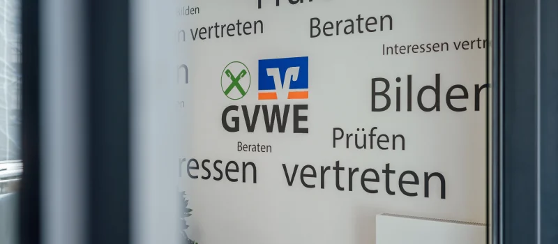 Blick durch eine Glastür auf eine Wand mit GVWE-Logo und Begriffen wie Prüfen, Beraten, Bilden und Interessen vertreten. Das Bild begleitet die Seite zu den Mitgliedsgenossenschaften des GVWE.