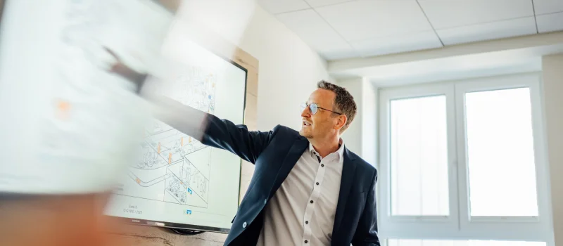 Ein Mann in Geschäftskleidung zeigt in einem Besprechungsraum auf eine digitale Darstellung. Das Bild steht für die strategische Nachhaltigkeitsberatung von Genossenschaftsbanken.
