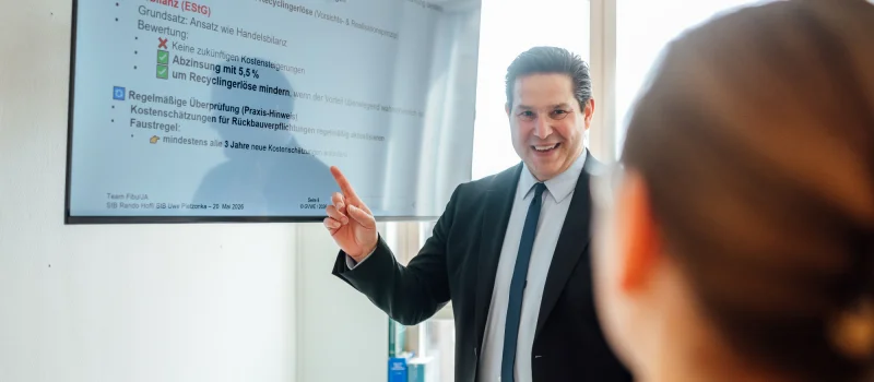 Ein Berater steht vor einem Bildschirm mit steuerlichen Stichpunkten und zeigt auf eine Präsentation. Das Bild steht für steuerliche Orientierung und fachliche Steuerberatung beim Genossenschaftsverband Weser-Ems.