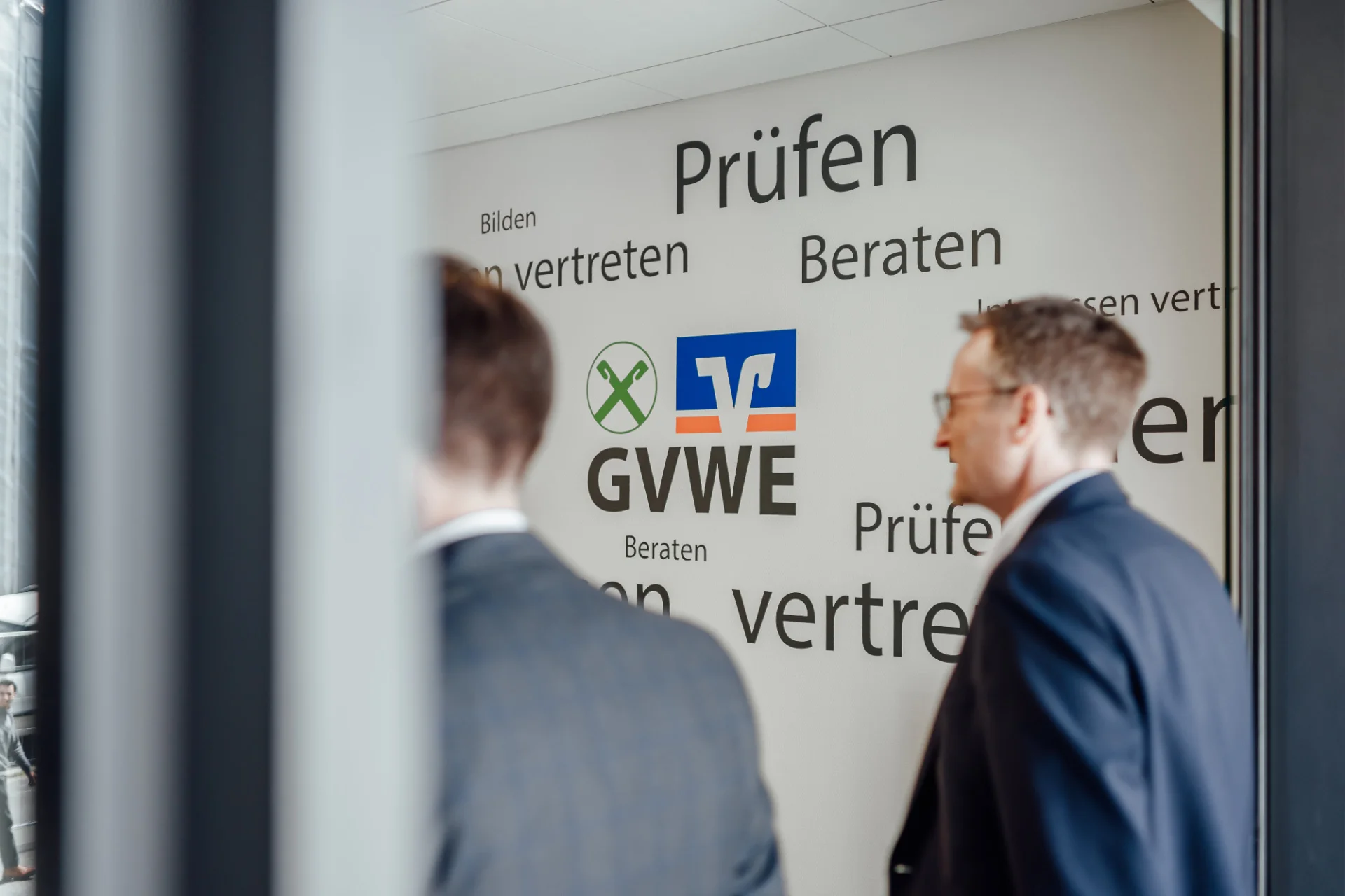 Zwei Personen stehen vor einer Wand mit GVWE-Logo und Begriffen wie Prüfen, Beraten, Bilden und Interessen vertreten. Das Bild begleitet den Abschnitt zu den Transparenzberichten und zur Struktur und Organisation des Verbands.