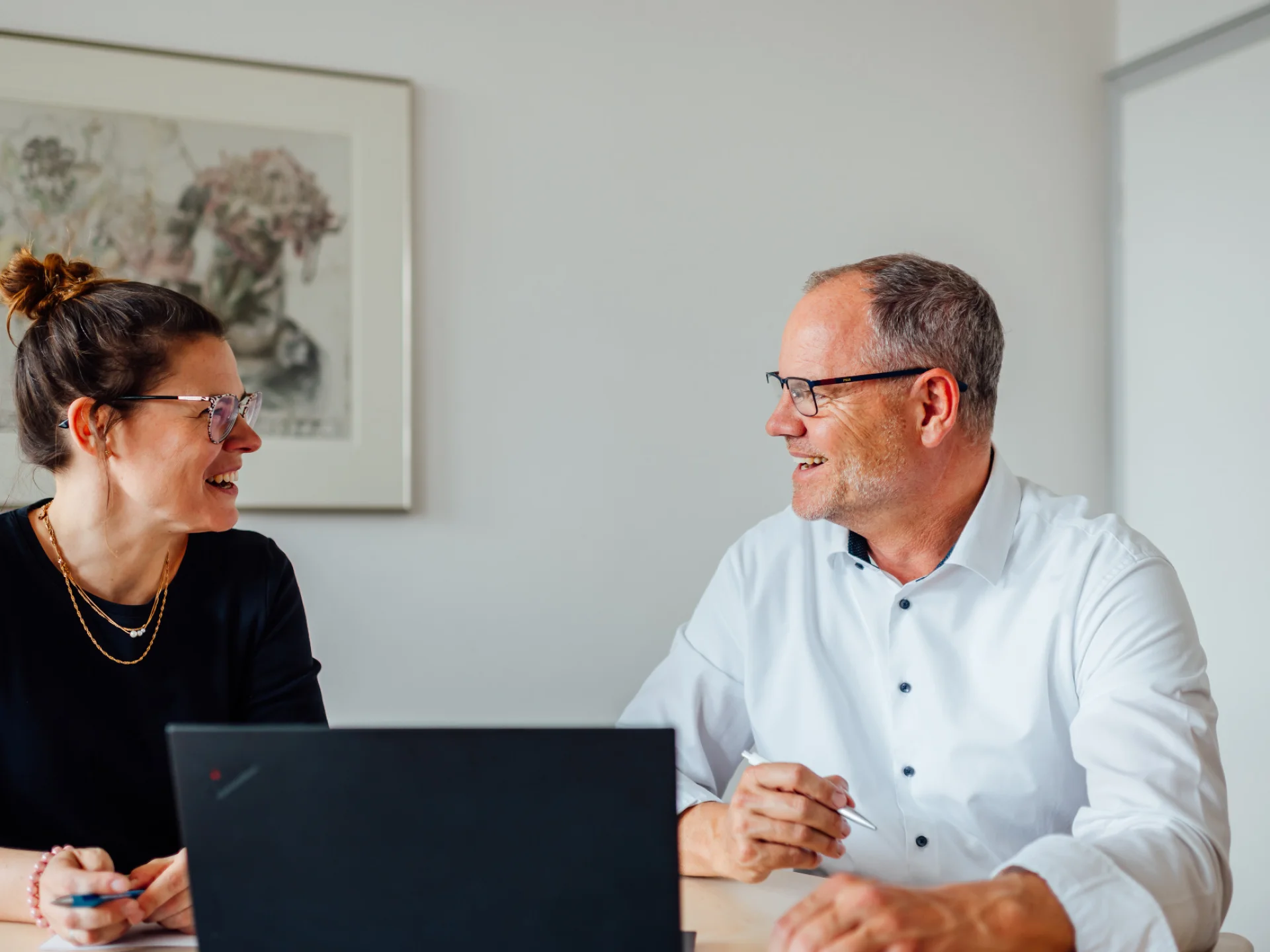 Zwei Personen sitzen an einem Tisch mit Laptop und sprechen miteinander. Das Bild steht für praxisnahen Austausch und Unternehmensberatung für Waren-, Vieh- und Dienstleistungsgenossenschaften.