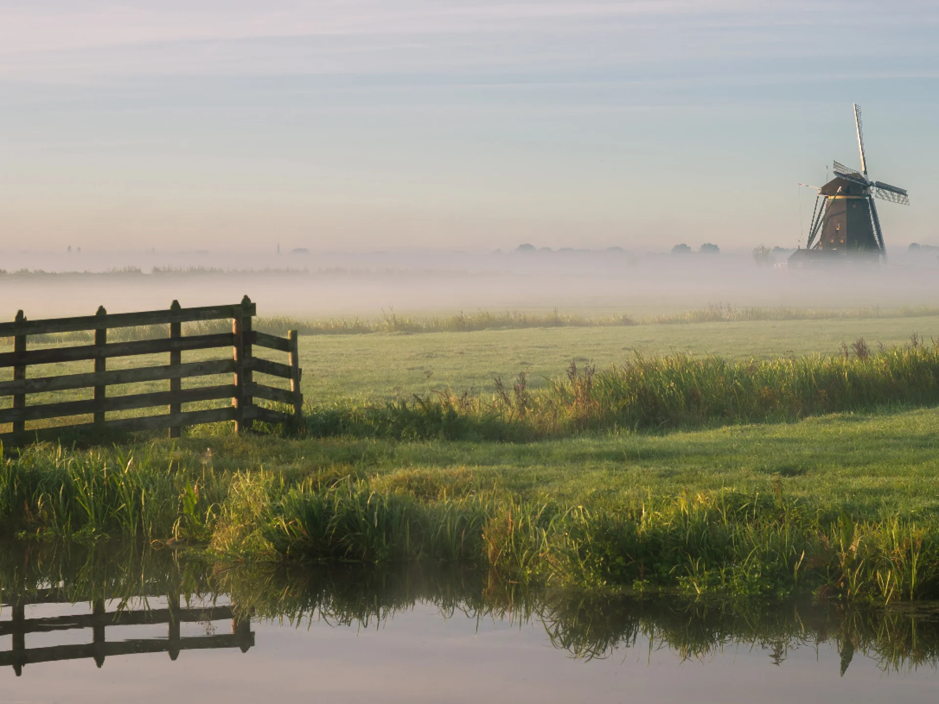 Eine weite Landschaft mit Wasserlauf, Wiese, Nebel und einer Windmühle im Hintergrund. Das Bild steht für die regionale Verwurzelung der Mitglieder des Genossenschaftsverbands Weser-Ems im Norden.