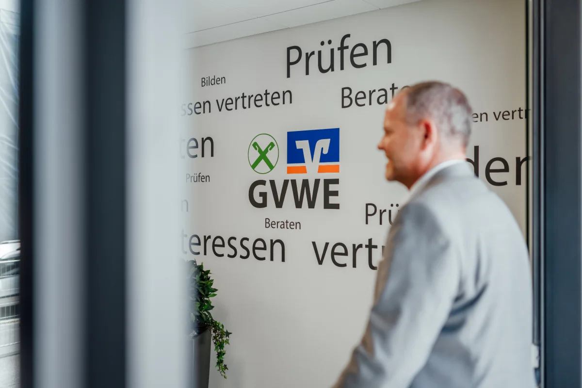 Eine Person steht vor einer Wand mit GVWE-Logo und Begriffen wie Prüfen, Beraten, Bilden und Interessen vertreten. Das Bild steht für die Satzung und die Aufgaben des Genossenschaftsverbands Weser-Ems.