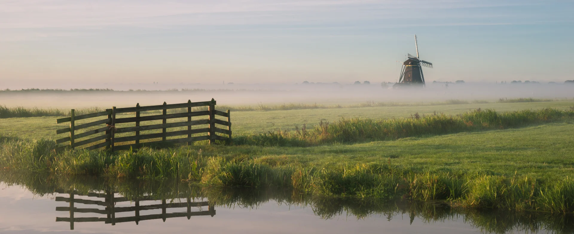 Eine weite Landschaft mit Wasserlauf, Wiese, Nebel und einer Windmühle im Hintergrund. Das Bild steht für die regionale Verwurzelung der Mitglieder des Genossenschaftsverbands Weser-Ems im Norden.